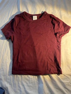 Vinröd t-shirt från Gina Tricot - En enkel och stilren vinröd t-shirt från Gina Tricot. T-shirten har rund halsringning och korta ärmar. Tillverkad i mjuk bomull som känns skön mot huden. Perfekt basplagg att matcha med jeans eller kjol.