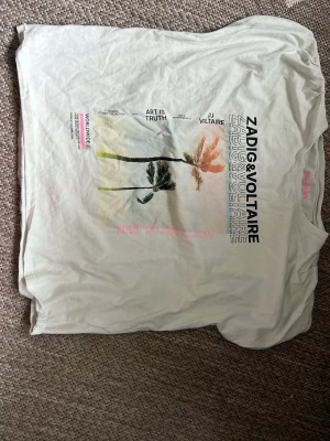Vit Zadig & Voltaire t-shirt med tryck - Säljer en vit t-shirt från Zadig & Voltaire med coolt tryck av palmer och text i svart, rosa och orange på framsidan. T-shirten har klassisk passform och korta ärmar. Perfekt till jeans eller shorts för en avslappnad look.
