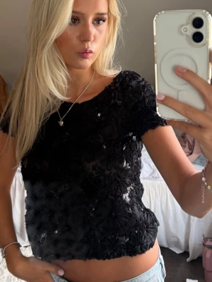 Svart paljett topp - Säljer en svart croppad topp med korta ärmar, täckt av paljetter och blommiga detaljer. Toppen har en lätt glittrig look och är perfekt för dig som vill sticka ut. Materialet känns syntetiskt och toppen har en figurnära passform.