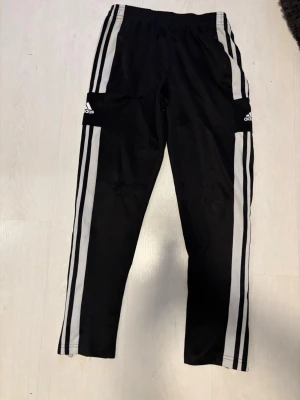 Adidas byxor  - Säljer ett par svarta mjukisbyxor från Adidas med klassiska vita ränder längs benen och elastisk midja. Byxorna har en avslappnad passform och är perfekta för chill dagar. Materialet är troligtvis polyester, vilket ger en sportig känsla.