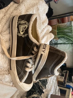Bruna sneakers från Roxy med foder - Bruna sneakers från Roxy med vitt foder och vita detaljer. Skorna har en vit sula med brun rand, vita skosnören och ett tryckt Roxy-logotyp på sidan. Utsidan är i mockaliknande material och insidan är fodrad för extra komfort.