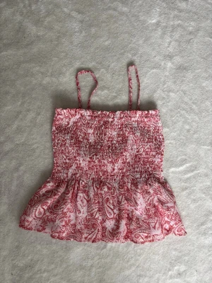 Rosa och vit smockad linnetopp - Supersöt linnetopp med smockad överdel och tunna axelband. Toppen är vit med rosa paisleymönster och har en lätt volang nedtill. Perfekt för varma dagar och ger en fräsch och ungdomlig look.