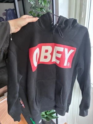 Svart OBEY hoodie med logga - Svart hoodie från OBEY med stor vit och röd logga på bröstet. Klassisk känguruficka framtill och justerbar huva med snören. Tillverkad i mjukt sweatshirtmaterial som passar perfekt för en avslappnad streetwear-look.