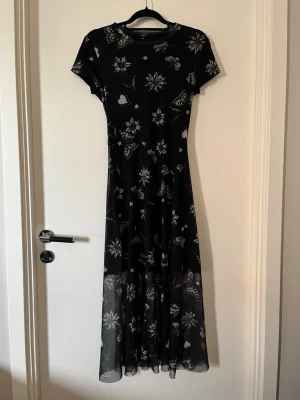Svart långklänning med fjärilar - Svart långklänning med korta ärmar och ett vitt mönster av fjärilar, blommor, hjärtan och stjärnor. Klänningen har en figurnära passform upptill och faller snyggt längs kroppen. Perfekt för dig som gillar unika prints och en stilren look.