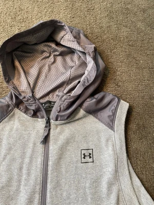 Under Armour Vest - Sportig väst från Under Armour, perfekt för träning eller avslappnad stil.  ✅ Grå melerad kroppstyg i mjuk fleece-liknande kvalitet ✅ Kontrasterande huva och axelpartier i tunnare, vindtätt material ✅ Nätfoder inuti huvan för andningsbarhet ✅ Halv dragkedja fram med Under Armour-logga ✅ Sido dragkedja för ventilation ✅ Perfekt lager för träningspass eller till en tröja underst  Skick: Mycket bra Storlek: M  En skön och funktionell väst som är lätt att styla, både till gymmet och vardagen. Hör av