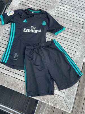 Real Madrid svart matchställ Adidas - Säljer ett svart Real Madrid matchställ från Adidas med turkosa detaljer. Setet består av en kortärmad tröja och shorts, båda med klassiska Adidas-ränder och klubbmärke. Materialet är lätt och andas, perfekt för fotbollsträning eller match. Defekt: Litet hål vid halsen men inget som ögat kan se på håll‼️‼️(se på sista bilden)