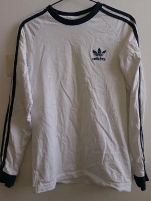 Vit långärmad Adidas tröja - Vit långärmad tröja från Adidas med svarta detaljer. Klassiska tre svarta ränder längs ärmarna och svart Adidas-logga på bröstet. Rund halsringning och muddar i svart. Perfekt för en sportig och avslappnad stil. Använd knappt