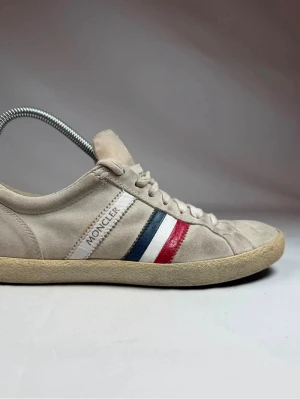 Beige Moncler sneakers med ränder - Snygga beige sneakers från Moncler i mocka med klassiska ränder i blått, vitt och rött på sidan. Skorna har lågt skaft, beige snörning och en beige sula i gummi. Perfekta för dig som gillar stilrena och sportiga detaljer. Storlek 40 men passar 41