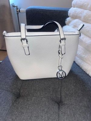 Vit handväska med metallcharm - Snygg vit handväska i konstläder med två justerbara handtag och en dekorativ metallcharm med bokstaven M. Väskan har en dragkedja upptill och en praktisk ficka med dragkedja på baksidan. Perfekt för dig som gillar stilrena accessoarer.