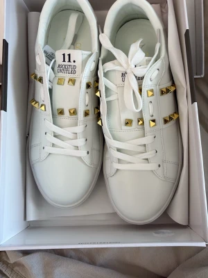 Vita Valentino Rockstud sneakers - Snygga vita sneakers från Valentino med guldiga nitar längs sidorna och på plösen. Skorna har klassisk låg profil, vita skosnören och är tillverkade i slätt läder. Perfekta för dig som vill sticka ut med exklusiva detaljer och modern design.