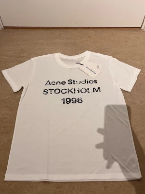 Vit Acne Studios t-shirt med tryck - Vit t-shirt från Acne Studios med svart tryck på bröstet där det står 'Acne Studios STOCKHOLM 1996'. Klassisk passform och rund hals. Perfekt för dig som gillar stilrena plagg med en twist.