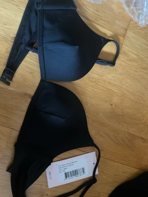 Svart Illusions Bikini Bra från Nelly - Snygg svart Illusions Bikini Bra från Nelly med släta kupor och justerbara axelband. Bh:n har enkel knäppning bak och är tillverkad i ett mjukt, stretchigt material som ger en bekväm passform. Perfekt för dig som vill ha en stilren och klassisk look.