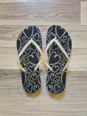 Svarta flipflops med hjärtmönster - Snygga svarta flipflops med vita hjärtan och vita remmar. Perfekta för sommaren och strandhäng. Mjukt syntetmaterial som är bekvämt att gå i och lätt att ta på sig.