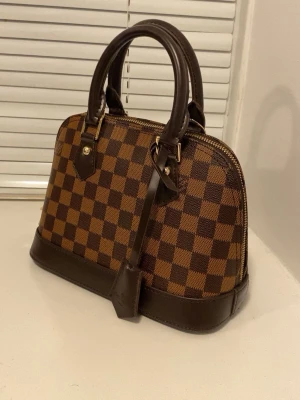 Brun rutig handväska i läderlook - Snygg handväska i klassiskt brunt rutigt mönster med mörkbruna läderdetaljer och guldfärgade metalldelar. Väskan har två handtag, dragkedja upptill och en elegant formad botten. Perfekt accessoar för att lyfta din outfit.