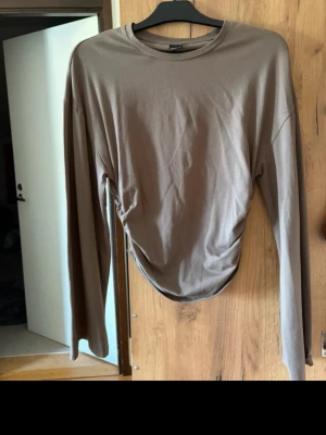 Brun långärmad topp med rynk - Snygg brun långärmad topp med rund hals och rynkade detaljer på sidorna. Tillverkad i ett mjukt och stretchigt material som sitter bekvämt på kroppen. Perfekt att styla med jeans eller kjol.