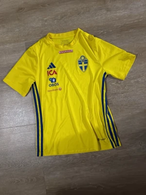 Svensk landslagströja Adidas gul - Gul svensk landslagströja från Adidas med blå detaljer och flera sponsorer på bröstet. Tröjan har korta ärmar, rund hals och Sveriges landslagsemblem på vänster sida. Perfekt för fotbollsträning eller att heja på Sverige i.