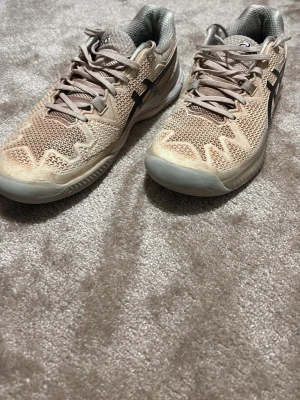 Beige sportskor från Asics - Säljer ett par beige sportskor från Asics med svart logga på sidorna. Skorna har mesh-ovandel för bra ventilation och slitstark sula med fiskbensmönster för extra grepp. Perfekta för träning eller sportaktiviteter.