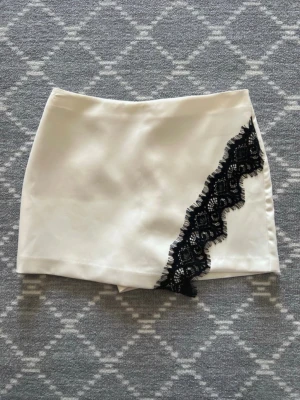 Populär Zara Boxkjol med svart spetsdetalj - Säljer en snygg vit kortkjol i satinliknande material med en diagonal svart spetsdetalj framtill. Kjolen har en clean och modern look med en unik touch av spets som gör den extra speciell. Perfekt för dig som vill sticka ut lite extra.