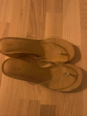 Beiga kilklackssandaler från Zara - Snygga beiga sandaler från Zara med kilklack och transparenta remmar. Skorna har öppen tå och en stilren design som passar perfekt till sommarens outfits. Materialet är en mix av syntet och plast, vilket ger en modern look.
