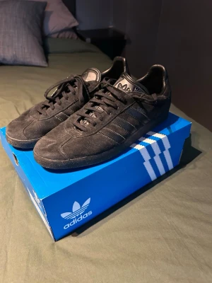 Svarta Adidas sneakers i mocka - Säljer ett par svarta Adidas sneakers i mocka med klassiska tre ränder på sidan och svart sula. Skorna har lågt skaft och snörning framtill. Perfekta för dig som gillar stilrena och tidlösa sneakers. Ingen box ingår