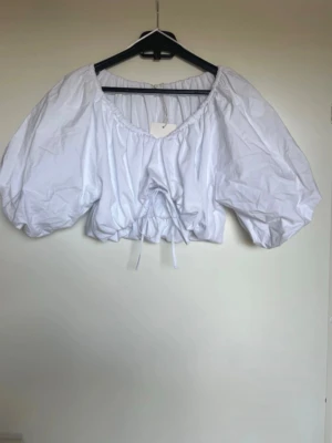 Populär Zara Vit croppad blus med puffärm - Säljer en populär viral vit croppad blus med stora puffärmar och snörning framtill. Toppen har en rundad halsringning och är tillverkad i ett lätt bomullstyg. Perfekt för dig som vill ha en trendig och luftig topp till sommaren. Passar både S och M