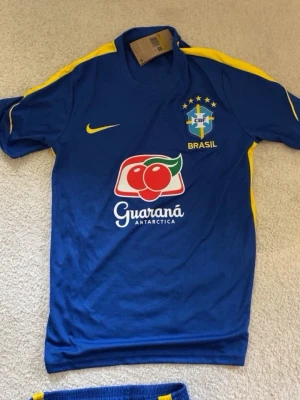 Blå Brasilien fotbollströja Nike - Säljer en blå Brasilien fotbollströja från Nike med gula detaljer och officiella loggor. Tröjan har CBF-emblemet, Guaraná Antarctica-tryck på framsidan och BRASA-tryck på ryggen. Perfekt för dig som älskar fotboll och vill stötta Brasilien.