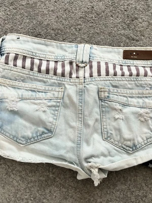 Ljusblå lågmidjade jeansshorts - Säljer ett par lågmidjade ljusblå jeansshorts med slitna detaljer, råa kanter och coolt tryck av vita stjärnor och ränder. Shortsen har fem fickor och låg midja. Perfekta för sommaren och ger en avslappnad vibe. Köpte på en secondhand i Polen, där av svårt att få tag i. Midjemått 39 cm💕