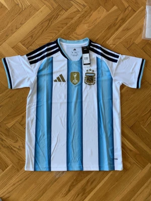 Argentina fotbollströja Adidas - Säljer en officiell Argentina fotbollströja från Adidas med klassiska blå och vita ränder. Tröjan har svarta detaljer på axlarna, FIFA- och AFA-emblem på bröstet samt rund hals. Tillverkad i lätt och ventilerande material, perfekt för träning eller match.