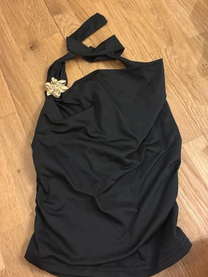 Svart halterneck tröja  med blomdetalj - Snygg svart tröja i halterneck-modell med dekorativ guldfärgad blomma vid axeln. Tillverkad i stretchigt material av 95% polyester och 5% elastan. 