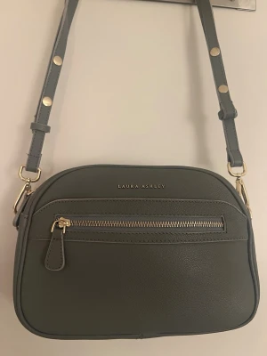 Laura Ashley grå axelremsväska i skinn - Grå axelremsväska i skinn från Laura Ashley med justerbar axelrem och guldfärgade detaljer. Väskan har ett fack med dragkedja på framsidan. Måtten är 25/ 27/8 cm. Fint skick  Jag säljer fler fina plagg, väskor, skor, accessoarer, parfymer och smycken. Se mina övriga annonser. Jag samfraktar förstås .