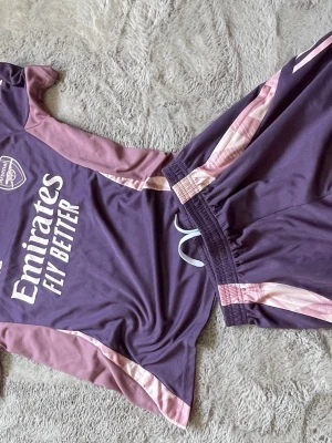 Lila Arsenal träningsset från Adidas - Säljer ett lila Arsenal träningsset från Adidas med tröja och shorts. Setet har ljusrosa detaljer och tryck med 'Emirates Fly Better' på tröjan. Perfekt för fotbollsträning eller sportiga dagar. Materialet är lätt och andas bra.