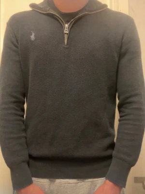 Svart halvzip tröja Polo Ralph Lauren - Svart stickad tröja från Polo Ralph Lauren med halvzip och hög krage. Tröjan har lång ärm, ribbade muddar och den klassiska broderade loggan på bröstet. Perfekt för lager på lager och en clean look.