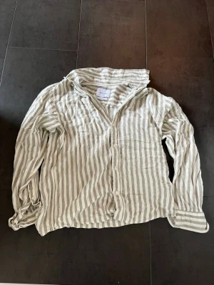 Randig linneskjorta från Vailent XS - Säljer en beige och vit randig skjorta från Vailent i linnemix. Skjortan har klassisk krage, knäppning framtill och långa ärmar. Perfekt för en avslappnad och stilren look.