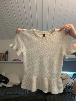 Vit stickad topp med volang - Supersöt vit stickad topp med korta ärmar och volangkant nedtill. Toppen har en rund halsringning och ribbade detaljer vid ärmslut och nederkant. Perfekt att styla med jeans eller kjol för en trendig look.