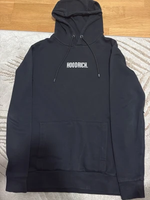 Svart hoodie från Hoodrich - Snygg svart hoodie från Hoodrich med vit logga tryckt på bröstet. Klassisk modell med huva och dragsnören samt stor magficka framtill. Tillverkad i mjukt och bekvämt material, perfekt för en avslappnad streetwear-look.