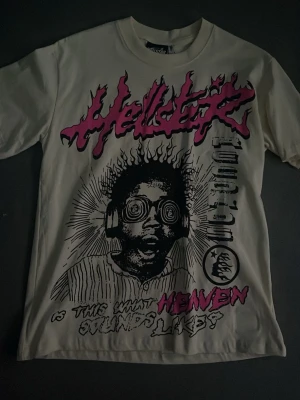 Vit Hellert Records t-shirt med tryck - Säljer en vit t-shirt med stort grafiskt tryck från Hellert Records. Framsidan har ett coolt motiv i svart och rosa med text och illustration, baksidan har Hellert Records-loggan. Klassisk passform och korta ärmar, perfekt för dig som gillar streetwear.