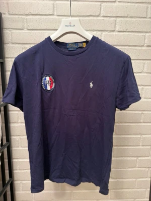 Mörkblå t-shirt från Polo Ralph Lauren - Snygg mörkblå t-shirt från Polo Ralph Lauren med klassisk broderad logga på bröstet och ett FR-märke på vänster sida. T-shirten har rund hals och korta ärmar, perfekt till jeans eller shorts.