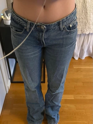 Blå raka jeans med låg midja - Ett par klassiska blå jeans med låg midja och raka ben. Jeansen har fem fickor och stängs med dragkedja och knapp. Materialet är denim i 100% bomull och färgen är en tvättad blå nyans. Perfekta för en avslappnad och trendig look.