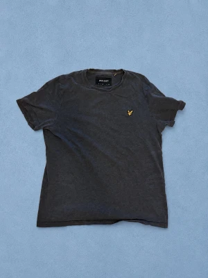 Lyle & Scott t-shirt  - Snygg mörkgrå t-shirt med en gul broderad logga på bröstet. Klassisk rund halsringning och korta ärmar. Tillverkad i mjuk bomull som känns skön mot huden. Perfekt till jeans eller shorts för en avslappnad look.