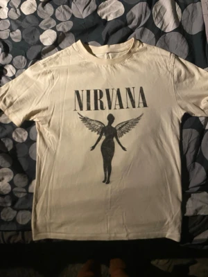 Vit Nirvana t-shirt med tryck - Säljer en vit t-shirt med klassiskt Nirvana-tryck framtill. T-shirten har korta ärmar och rund hals. Materialet känns som bomull och trycket visar en siluett med vingar under bandnamnet. Perfekt för dig som gillar bandtröjor och vill ha en stilren look.