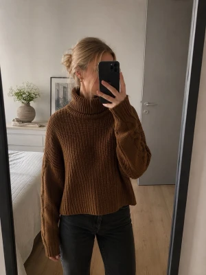 Brun stickad polotröja - Mysig brun stickad polotröja med grovstickad struktur och flätmönster på ärmarna. Tröjan har en hög, bred polokrage och avslappnad passform men är kort i modellen. 