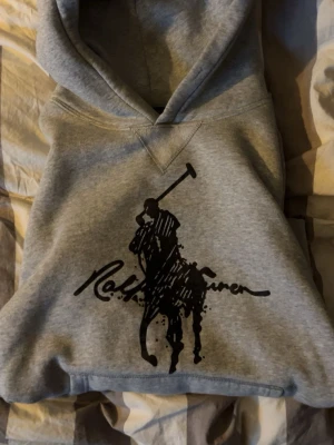 Grå hoodie från Polo Ralph Lauren - Snygg grå hoodie från Polo Ralph Lauren med stor svart logga och polospelare på bröstet. Klassisk huva och ribbade muddar. Tillverkad i mjukt material som är skönt att bära. Perfekt för en avslappnad och trendig look. Storlek L men passar även dig om du har M. Vid frågor tveka inte på att höra av dig!🤝