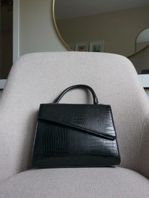 Svart handväska - Svart handväska i konstläder med handtag och unik asymmetrisk lockdesign. Väskan kommer tillsammans med ett av och påtagbart axelband.