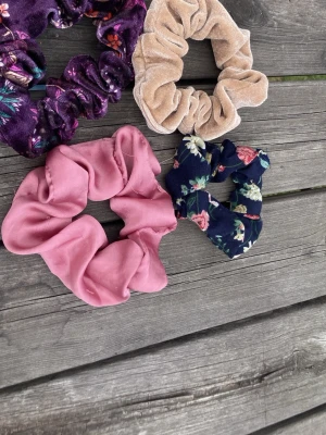 Fyra scrunchies i olika färger - Säljer ett set med fyra scrunchies i olika material och färger: en lila med blommönster i sammet, en beige i mjuk frotté, en rosa i sammetsliknande tyg och en marinblå med blommigt mönster. 