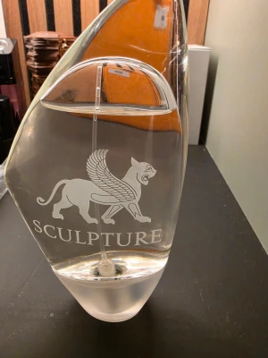 Sculpture parfymflaska med lejonmotiv - Unik parfymflaska i genomskinligt glas med ett stilrent lejon med vingar tryckt på framsidan och texten SCULPTURE. Flaskan har en modern, asymmetrisk form och en transparent spraydel. Perfekt som snygg detalj på hyllan.