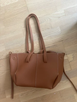 Brun tote handväska i konstläder - Snygg brun tote handväska i konstläder med två långa axelband och vita kontrastsömmar. Väskan har en enkel, stilren design och rymligt huvudfack. Perfekt för dig som vill ha en klassisk och praktisk väska till vardagen.