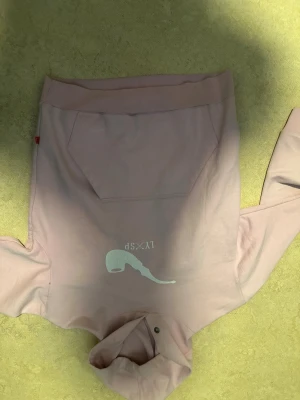 Ljusrosa hoodie med tryck - Säljer en ljusrosa hoodie med huva och magficka. På framsidan finns ett vitt tryck med texten 'LVXS' och en grafisk illustration. Hoodien har ribbade muddar och en avslappnad passform. Perfekt för en chill och stilren look.