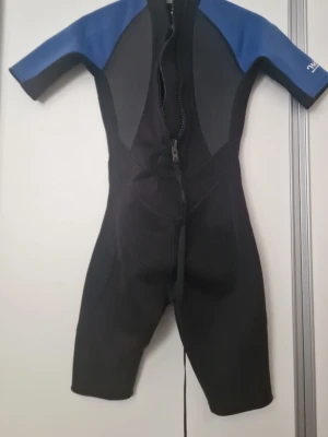 Svart och blå kort våtdräkt Wetnfun - Kortärmad våtdräkt från Wetnfun i svart och blått. Dräkten har dragkedja fram och är tillverkad i ett stretchigt material, troligen neopren, som ger bra rörelsefrihet. Perfekt för strand och vattensport. Storlek 155