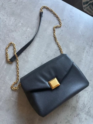 Svart axelväska med guldkedja - Snygg svart axelväska i slätt läder med guldfärgad kedja och fyrkantigt guldfärgat spänne framtill. Väskan har en stilren och modern design, perfekt att bära crossbody. Enkel att matcha med olika outfits.