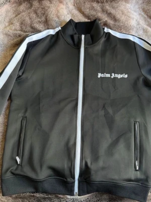 Svart track jacket från Palm Angels - Snygg svart track jacket från Palm Angels med vit dragkedja och vita ränder längs ärmarna. Jackan har ståkrage, två fickor med dragkedja och logga på bröstet. Perfekt för en sportig och trendig look. Vid snabb affär kan jag gå ner lite i pris 